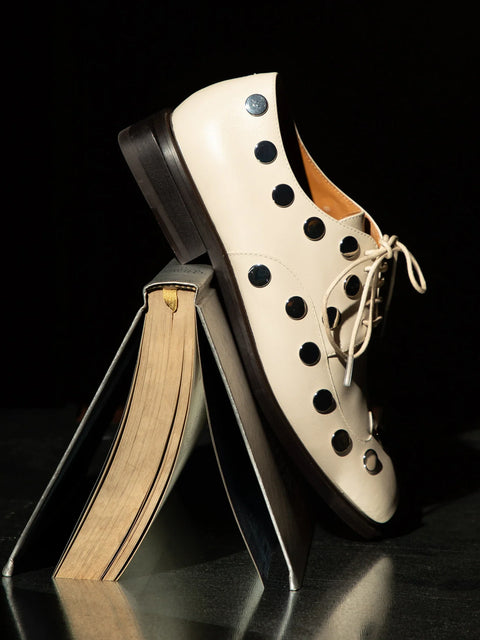 A cream-colored lace-up derby shoe with mirror stud, displayed on a stack of books against a black background.