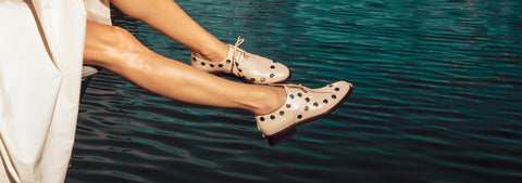 A person sitting by the water with their legs dangling over the edge, wearing beige lace-up derby shoes with studs.